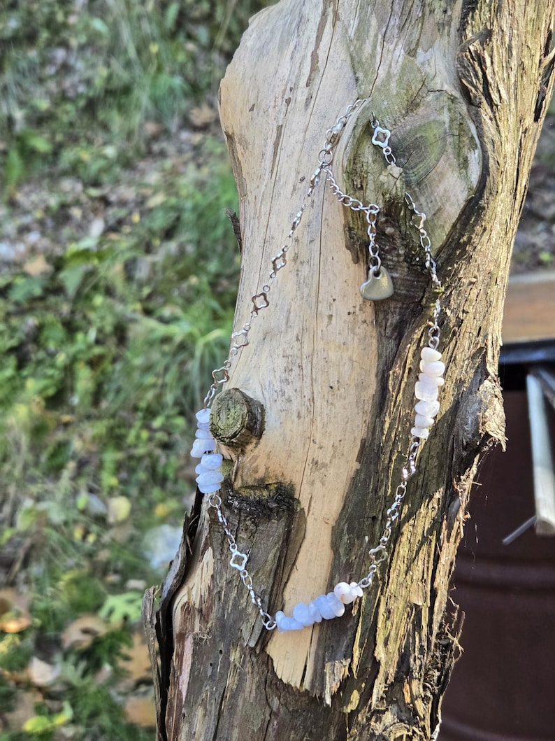 May include: A delicate silver necklace with a heart pendant and light blue gemstone beads. The necklace is draped over a weathered tree trunk, showcasing its intricate design and natural aesthetic. The necklace features a chain with small square-shaped links.