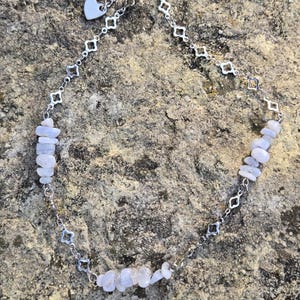 May include: A silver-toned necklace featuring a delicate chain with a heart-shaped charm and diamond-shaped links. The necklace is adorned with small, light blue gemstone chips, creating a subtle, natural aesthetic. The necklace is displayed on a textured, neutral-toned surface.