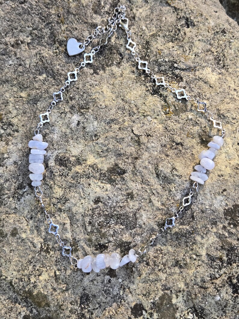 May include: A silver-toned necklace featuring a delicate chain with a heart-shaped charm and diamond-shaped links. The necklace is adorned with small, light blue gemstone chips, creating a subtle, natural aesthetic. The necklace is displayed on a textured, neutral-toned surface.