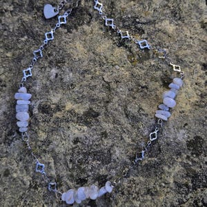May include: A delicate necklace featuring a silver chain with alternating geometric shapes and small blue-grey gemstone chips. A small heart charm dangles near the clasp. The necklace is displayed on a textured, grey stone surface.