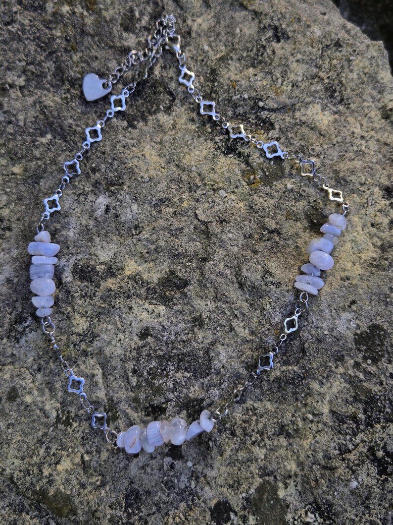 May include: A delicate necklace featuring a silver chain with alternating geometric shapes and small blue-grey gemstone chips. A small heart charm dangles near the clasp. The necklace is displayed on a textured, grey stone surface.