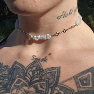 May include: A delicate choker necklace featuring small, light blue gemstones and silver-toned chain links. The necklace is worn around the neck. The word "Hold" is tattooed on the neck. The word "Speak" is tattooed on the chest.