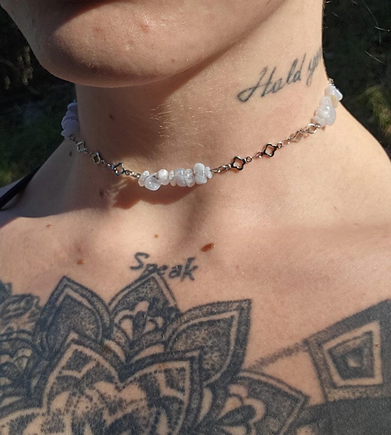 May include: A delicate choker necklace featuring small, light blue gemstones and silver-toned chain links. The necklace is worn around the neck. The word "Hold" is tattooed on the neck. The word "Speak" is tattooed on the chest.