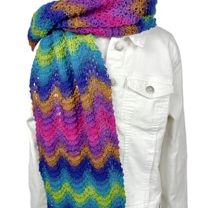 May include: A multi-colored, open-knit scarf featuring a wavy, chevron-like pattern in vibrant shades of pink, purple, blue, green, and gold. The scarf is draped over a crisp, white denim jacket with silver-toned metal buttons and chest pockets.