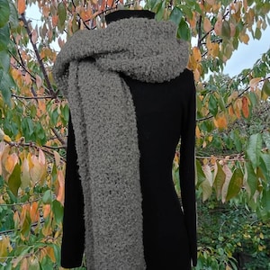 May include: A gray, textured scarf draped over a black garment. The scarf is made of a soft, fluffy material. The background features a tree with green, orange, and yellow leaves.