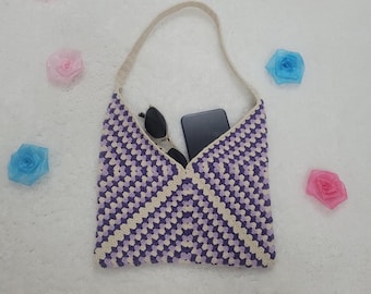 May include: A handmade crochet bag in a purple and cream geometric pattern. The bag contains a pair of sunglasses and a black smartphone. The bag has a cream-colored strap. Decorative floral accents surround the bag.