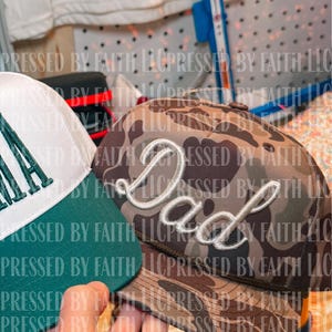 Op de afbeelding: Twee baseballpetten. Eén is wit en groen met het woord "MA" op de voorkant geborduurd. De andere is camouflage met het woord "Dad" in witte letters geborduurd. De petten worden tentoongesteld.