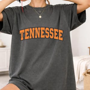 May include: A charcoal gray short-sleeve t-shirt featuring the word TENNESSEE printed in a bold, arched orange font with a black outline across the chest. The garment has a crew neckline and a relaxed, loose-fitting silhouette.