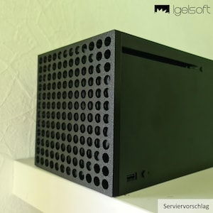 Könnte beinhalten: Eine schwarze Spielkonsole mit perforierter Frontplatte und einer soliden Seitenplatte. Die Konsole steht auf einem weißen Regal. Der Text "Serviervorschlag" befindet sich unten rechts.