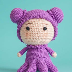 May include: A handmade crochet doll featuring a round beige face with black bead eyes and thin brown eyebrows. The doll wears a purple hooded outfit with two rounded ears on top, a ruffled collar, and a base shaped like multiple octopus tentacles.