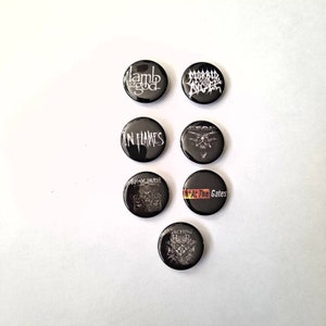 May include: A collection of seven black round buttons featuring white and red text and graphics. The buttons display band names such as "Lamb of God", "Morbid Angel", "In Flames", "At the Gates", and "Machine Head".
