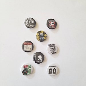 May include: A collection of round, black and white punk rock buttons with various band logos and graphics. The buttons feature text like "The Exploited", "GBH", and "Sub Humans". The buttons are scattered on a white surface.