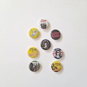 May include: A collection of eight round pinback buttons featuring band logos and graphics. The buttons are in various colors, including yellow, black, white, and green. Some buttons display text such as "The Clash" and "Sex Pistols."