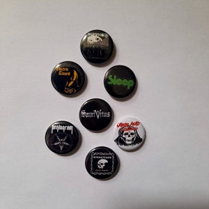 May include: A collection of seven black round buttons with various band logos. The buttons feature logos for bands like Sunn O))), Electric Wizard, Sleep, Saint Vitus, Pentagram, Eyehategod, and Uncle Acid & the Deadbeats. The buttons are arranged on a white surface.