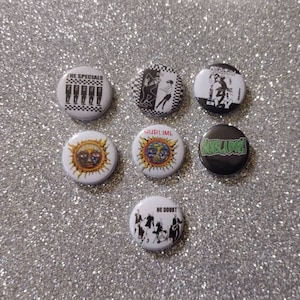 May include: A collection of seven round pinback buttons with various designs. The buttons feature band logos and graphics in black, white, green, and yellow. The text on the buttons includes "THE SPECIALS", "SUBLIME", and "NO DOUBT".