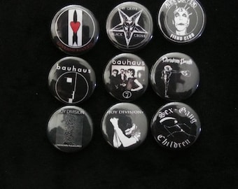 May include: Nine black and white button pins with various designs. The pins feature band names like "Bauhaus", "Joy Division", and "Alien Sex Fiend". Other pins have graphics of a heart, a cross, and a skeleton.