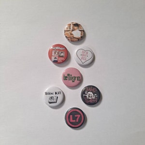 May include: A collection of seven round pinback buttons with various designs and text. The buttons feature images of a person reading, band logos, and text like "Jack Off Jill" and "Bikini Kill". The color palette includes red, pink, black, and white.