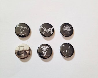 May include: Six black round buttons with white text and graphics. The buttons feature band logos, including "Bathory," "Mayhem," "Darkthrone," "Immortal," "Venom," and "Satyricon." The buttons are arranged on a white surface.