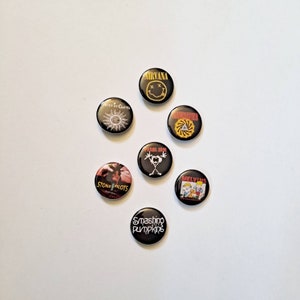 May include: A collection of seven black pinback buttons featuring band logos and names. Bands include Nirvana, Soundgarden, Pearl Jam, Alice in Chains, Stone Temple Pilots, Melvins, and Smashing Pumpkins. The buttons are round and vary in design.