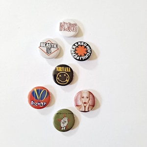 May include: A collection of seven round pinback buttons featuring band logos and images. Bands include Incubus, Beastie Boys, Red Hot Chili Peppers, Nirvana, No Doubt, and Rage Against the Machine. The buttons are in various colors, including red, black, and white.