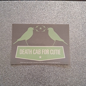 May include: A rectangular sticker with a gray background features two light green bird silhouettes facing each other. Above the birds are two lightning bolts and a star. The text "DEATH CAB FOR CUTIE" is displayed in a light green banner.