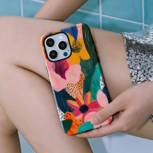 Könnte beinhalten: Bunte Handyhülle mit einem lebendigen Blumenmuster. Die Blumen sind rosa, orange, gelb und blau. Die Hülle hat eine glänzende Oberfläche und einen schwarzen Kameraausschnitt. Das Design umfasst grüne Blätter und schwarze gepunktete Akzente.