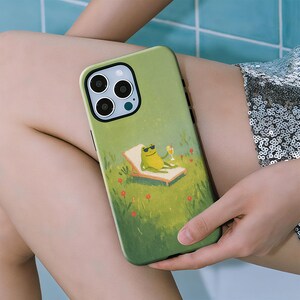 May include: A light green smartphone case featuring a printed illustration of a green frog wearing black sunglasses, lounging on a tan beach chair in a grassy field with small red flowers, while holding a yellow drink with a straw.