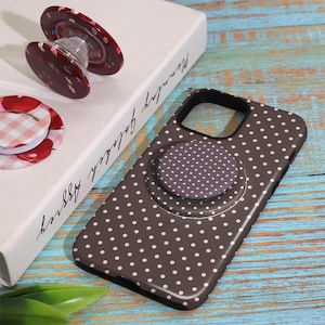 Pode incluir: Uma capa de telefone castanha com bolinhas brancas e uma pega pop-up a condizer. Pegas pop-up adicionais apresentam padr&otilde;es florais e xadrez. Os artigos s&atilde;o exibidos numa superf&iacute;cie de madeira azul e branca.