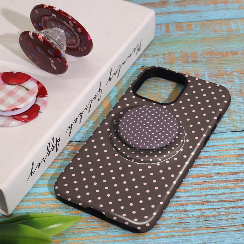 Pode incluir: Uma capa de telefone castanha com bolinhas brancas e uma pega pop-up a condizer. Pegas pop-up adicionais apresentam padr&otilde;es florais e xadrez. Os artigos s&atilde;o exibidos numa superf&iacute;cie de madeira azul e branca.