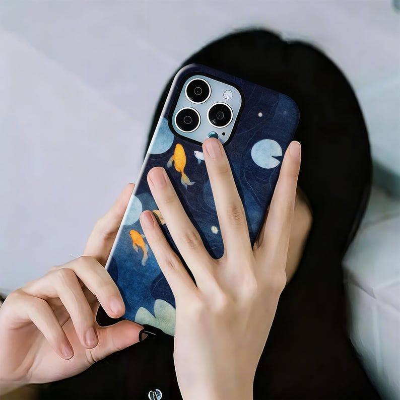 May include: A navy blue phone case featuring a koi pond design with orange fish, white lily pads, and a dark blue background. The case has a camera cutout with three lenses and a flash.