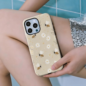 Puede incluir: Funda de teléfono beige con un patrón repetido de abejas de dibujos animados y margaritas blancas. La funda tiene un borde negro y un recorte para la cámara. El diseño incluye pequeños puntos amarillos y hojas verdes.