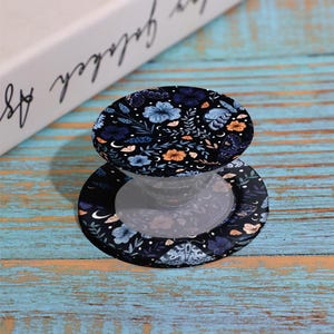 May include: A black phone grip with a floral design featuring blue and orange flowers, leaves, and crescent moons. The grip is on a blue and turquoise wooden surface. A book is in the background.