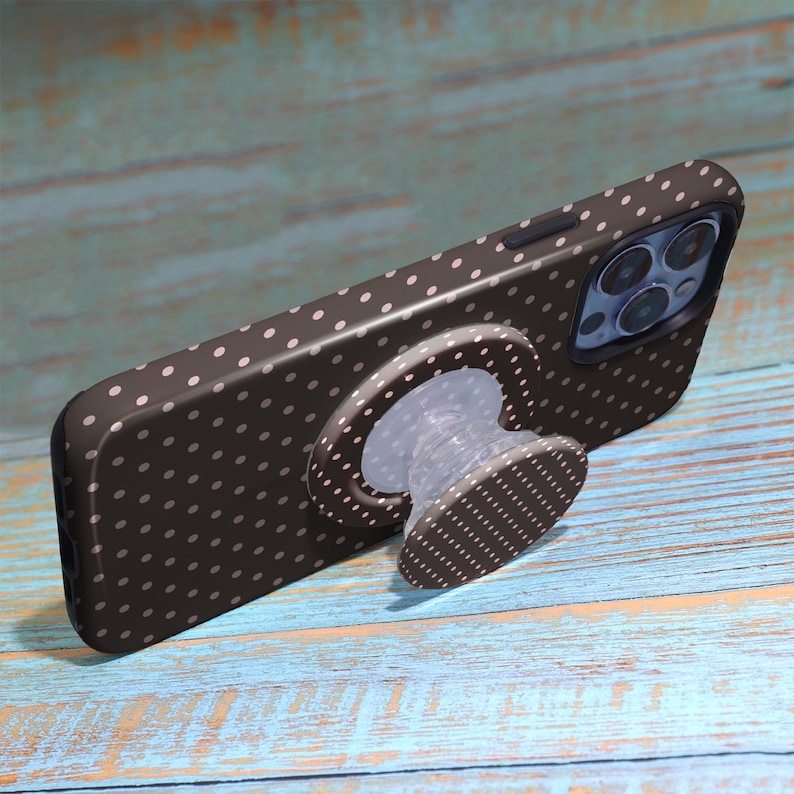 Pode incluir: Uma capa de telefone castanha com bolinhas brancas e um suporte a condizer. A capa do telefone est&aacute; numa superf&iacute;cie de madeira azul e castanha. O telefone tem v&aacute;rias lentes de c&acirc;mara.
