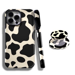 May include: A phone case, phone grip, and phone strap featuring a black and white cow print design. The phone case is on the left, with the phone grip and strap on the right. The phone case has a camera cutout.