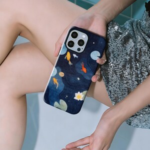 May include: A navy blue phone case featuring a koi pond design. The case has a dark blue background with yellow, orange, and white koi fish, lily pads, and a white lotus flower. The case has a glossy finish.