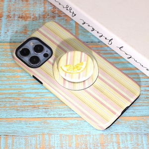 May include: A phone case with a yellow and pink striped pattern and a matching pop socket featuring a lemon slice design. The phone case is on a blue and teal wooden surface.