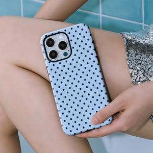 Op de afbeelding: Een lichtblauwe telefoonhoes met een herhalend patroon van kleine, donkerblauwe stippen. De hoes heeft een zwart camerakader en wordt in een hand gehouden. De telefoonhoes is een beschermend accessoire.