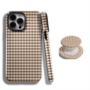 Puede incluir: Una funda de teléfono, un agarre para teléfono y una vista lateral de una funda de teléfono, todos con un patrón de cuadros marrones y blancos. La funda del teléfono tiene un recorte para la cámara y un borde negro. El agarre del teléfono es redondo y tiene un patrón a juego.