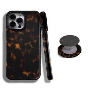 Puede incluir: Una funda y un agarre para teléfono con un patrón de carey. La funda es negra con un patrón marrón y amarillo. El agarre es redondo con un patrón a juego y una base transparente. La funda se muestra en posición vertical y de lado.