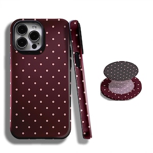 May include: A burgundy phone case and matching phone grip with a repeating pattern of small, light pink polka dots. The phone case is shown upright and on its side, with the grip next to it.