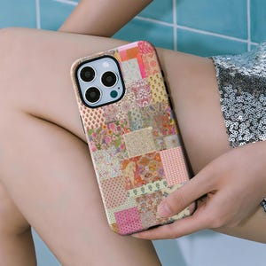 Op de afbeelding: Een smartphonehoesje met een veelkleurig patchworkontwerp met diverse bloemen-, geometrische en dierenpatronen in tinten roze, oranje, geel en groen. Het hoesje heeft een uitsparing voor een camerasysteem met drie lenzen aan de achterkant.