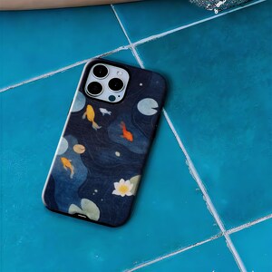 May include: A phone case featuring a dark blue design with koi fish, lily pads, and water plants. The case has a glossy finish and a camera cutout. The koi fish are orange and yellow.