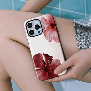 May include: A phone case with a cream-colored background featuring two hibiscus flower illustrations. One flower is a deep red, and the other is a lighter pink. The case has a black camera module and a black border.
