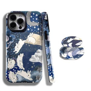May include: A phone case and matching pop socket featuring a whimsical night sky design. The case and pop socket showcase white rabbits, stars, clouds, and a crescent moon against a dark blue background.