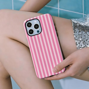 Op de afbeelding: Een roze en wit gestreept telefoonhoesje. De hoes wordt in de hand van een persoon gehouden en rust op hun been. De telefoonhoes heeft een zwarte rand rond de cameralenzen.