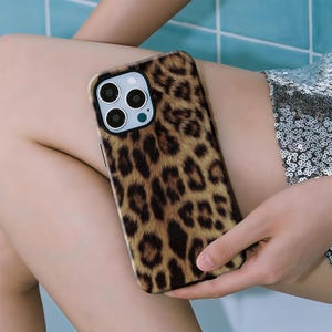 May include: A smartphone case with a leopard print design. The case features a black border and a brown and tan spotted pattern. The phone case is held in a person's hand.