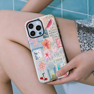 May include: A decorative phone case featuring a collage of beach-themed images. The case includes images of a turtle, cherries, flowers, and the words "Aloha, Howdy, Hello." The case is held by a person.
