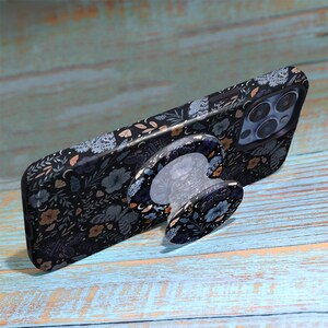 May include: A black smartphone case with a floral pattern, featuring blue, tan, and white flowers and leaves. The case has a matching phone grip attached. The phone case is on a light blue wooden surface.
