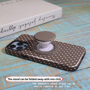 Pode incluir: Uma capa de telefone castanha com um padr&atilde;o de bolinhas brancas e um suporte de telefone pop-up a condizer. A pega pop-up &eacute; branca com um design de bolinhas castanhas. O texto na imagem diz: "O suporte pode ser dobrado com um clique."