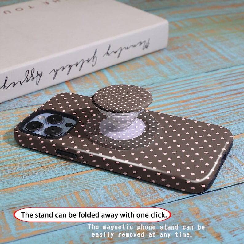 Pode incluir: Uma capa de telefone castanha com um padr&atilde;o de bolinhas brancas e um suporte de telefone pop-up a condizer. A pega pop-up &eacute; branca com um design de bolinhas castanhas. O texto na imagem diz: "O suporte pode ser dobrado com um clique."